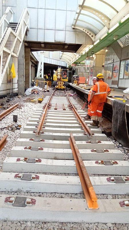 Track renewal works in progress at a station platform: new concrete sleepers and rails laid out on ballast, plant working under the canopy and an operative in orange high-visibility clothing on the platform edge.